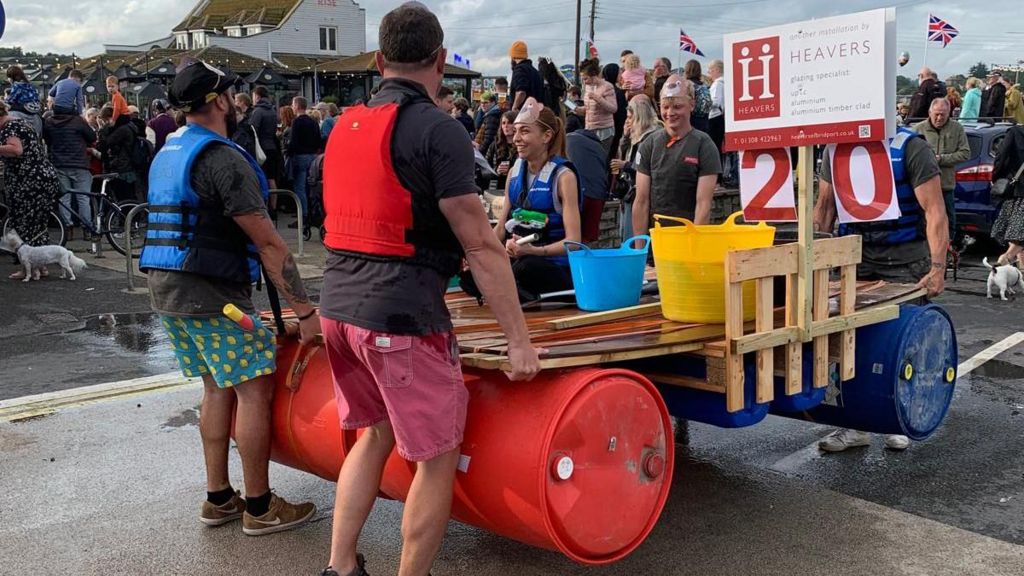 four people stood around a raft holding it up with one woman sat in the middle. All of them are wearing life jackets and are preparing to enter the water