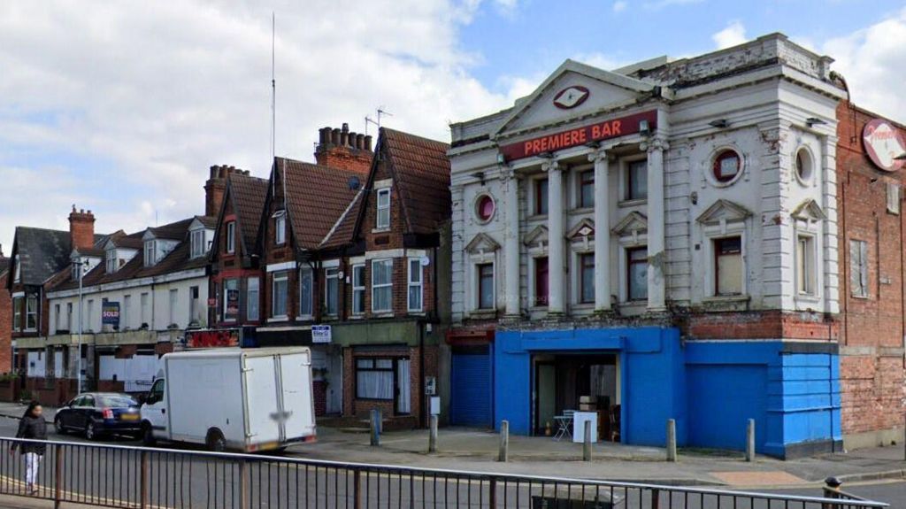 Derelict cinema with facade comprising pillars, white and blue brickwork and colonades. A red neon sign near the top of the building reads "Premiere Bar". The building stands in a street of other red-brick properties 