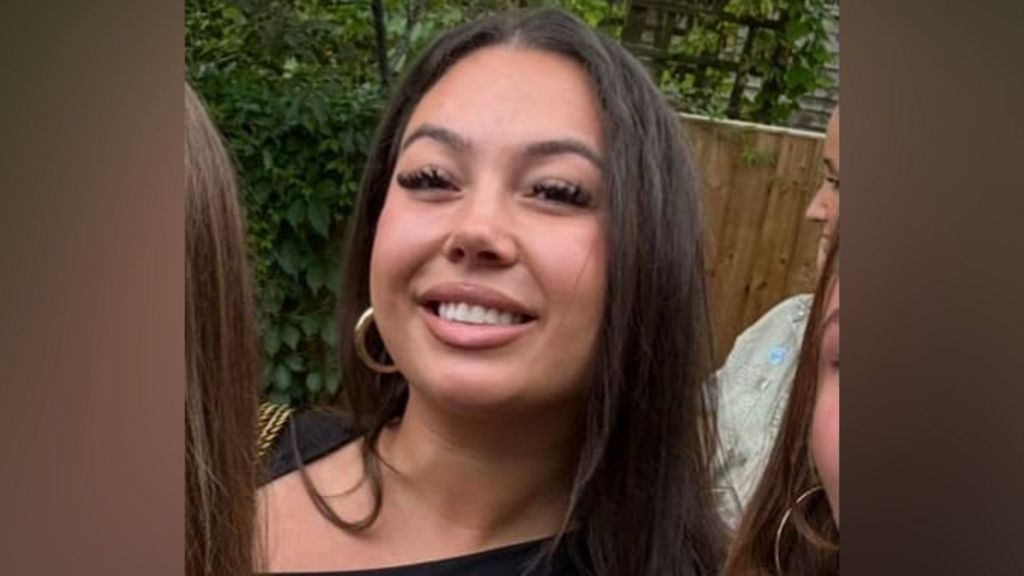 A close-up of a smiling young woman with long dark hair and gold hoop earrings, outdoors against a backdrop of green foliage and a wooden fence.
