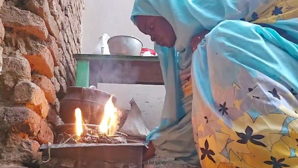 Manahel is crouching down by a fire, with cooking pots in the background. Her robe and headscarf are pale blue with yellow and dark brown flowers.