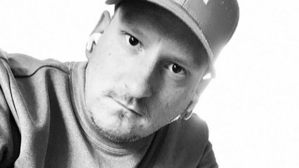 A black-and-white photo of a man wearing a cap and a top looking into to the camera. He is wearing white earbuds