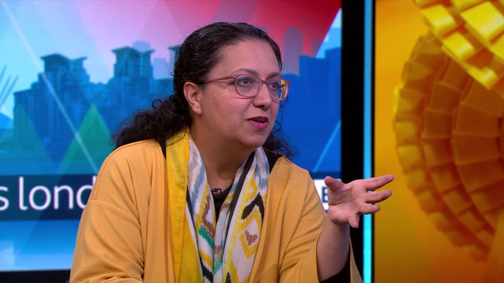 A woman wears a yellow cardigan and a patterned scarf along with pink framed glasses. She talks in a studio with a colourful background