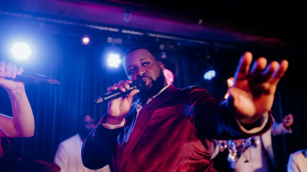 A male singer in a red velvet jacket looks into the crowd holding a microphone to his mouth. 