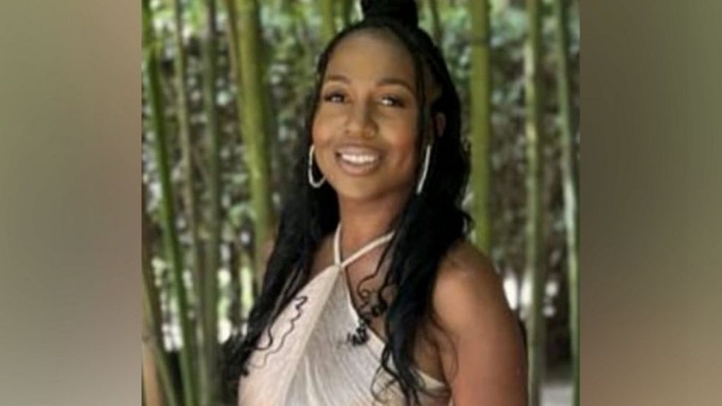 A young woman wearing gold jewellery smiles. She is standing in front of a forest.