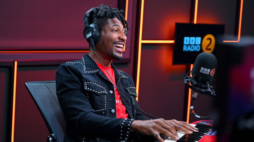 Jon Batiste sitting in a chair in the Radio 2 studio playing a small keyboard on the desk. He is wearing a black denim jacket with a red t-shirt.