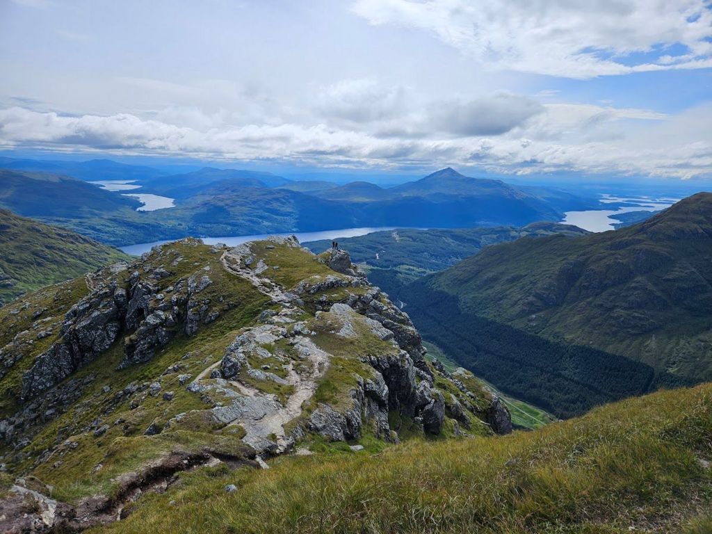 Panoramic view of grass and rock covered hills, surrounding a body of water below a white cloud covered sky. In the distance is the outline of two hill climbers.