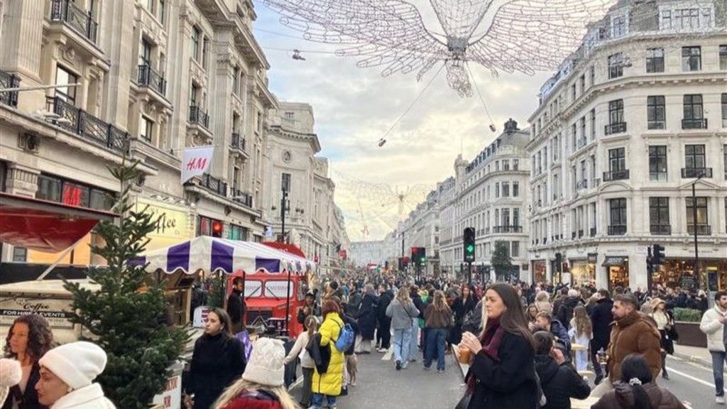 A picture of Regent Street during the festive celebrations. There are large crowds of people, a Christmas tree, a red bus parked on one side and the Christmas lights above the street. 