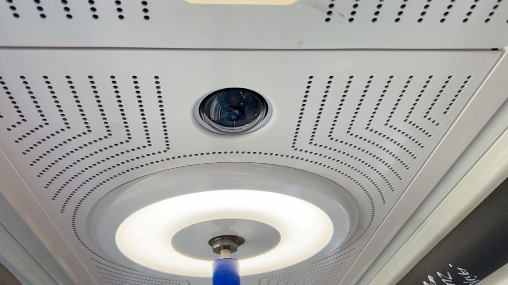 A camera is in the roof of the train and there is a circular light around a blue pole