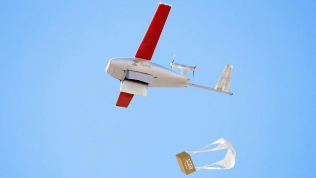 A drone flying in a blue sky with red wings carrying a parcel with a small parachute attached.