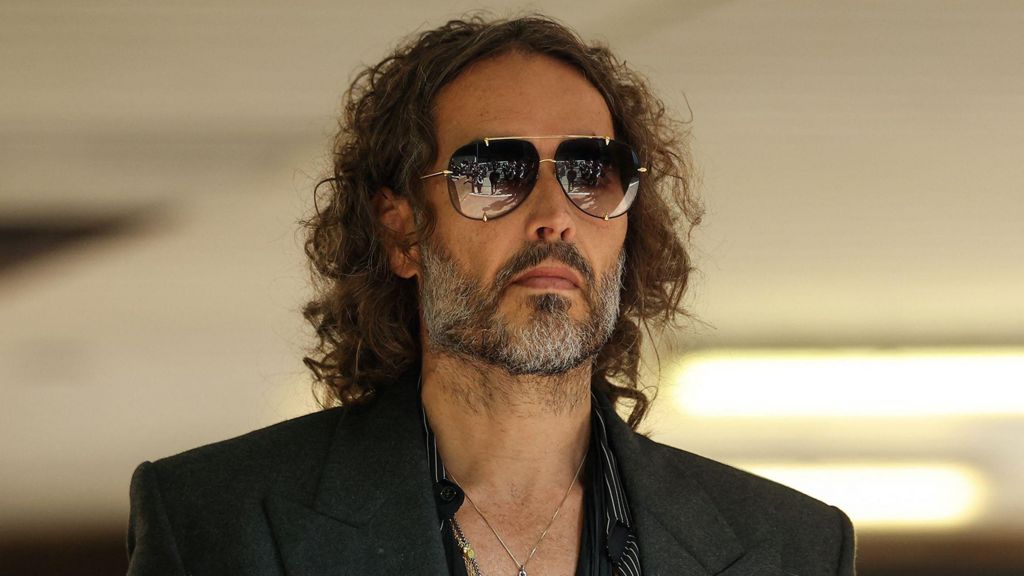 A headshot of Russell Brand who wears a suit and sunglasses.