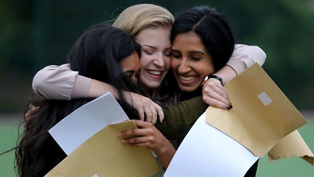 Three teenage girls smiling and hugging, holding their GCSE results in their hands.