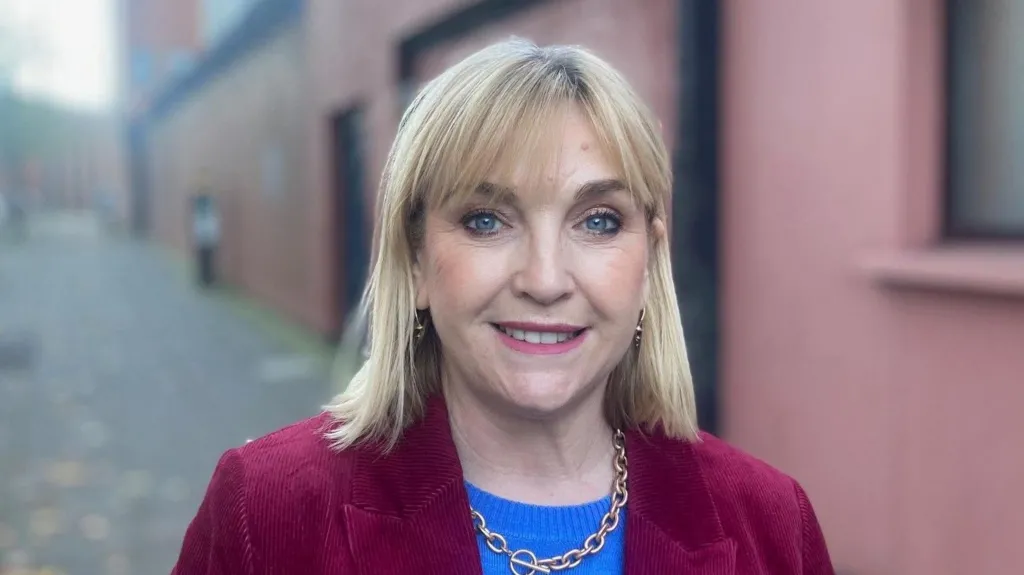 Celine McStravick has shoulder-length blonde hair and blue eyes. She is smiling at the camera. She is wearing a blue jumper and a maroon blazer. Behind her is a red wall.