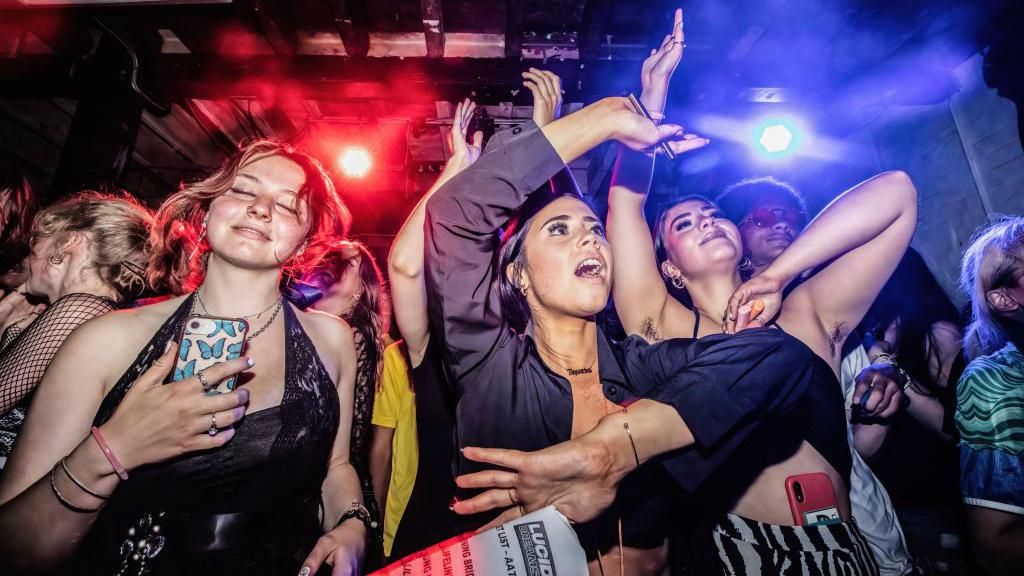 Three young women dancing in a Manchester nightclub with a crowd and flashing red and blue lights behind them