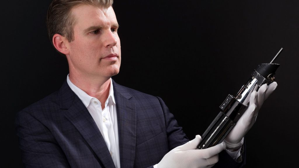 A man in a suit who is wearing white gloves is carefully holding and admiring a lightsaber prop against a black background