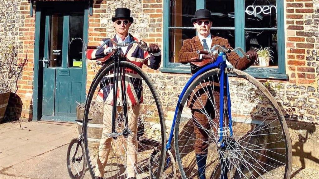 Two men, one dressed in a union flag jacket and a top hat and the other wearing a tweed suit and a bowler hat, standing outside a pub with their penny farthings