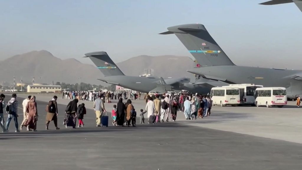 People carrying luggage walk across an airport tarmac towards large US military transport aircraft during an evacuation