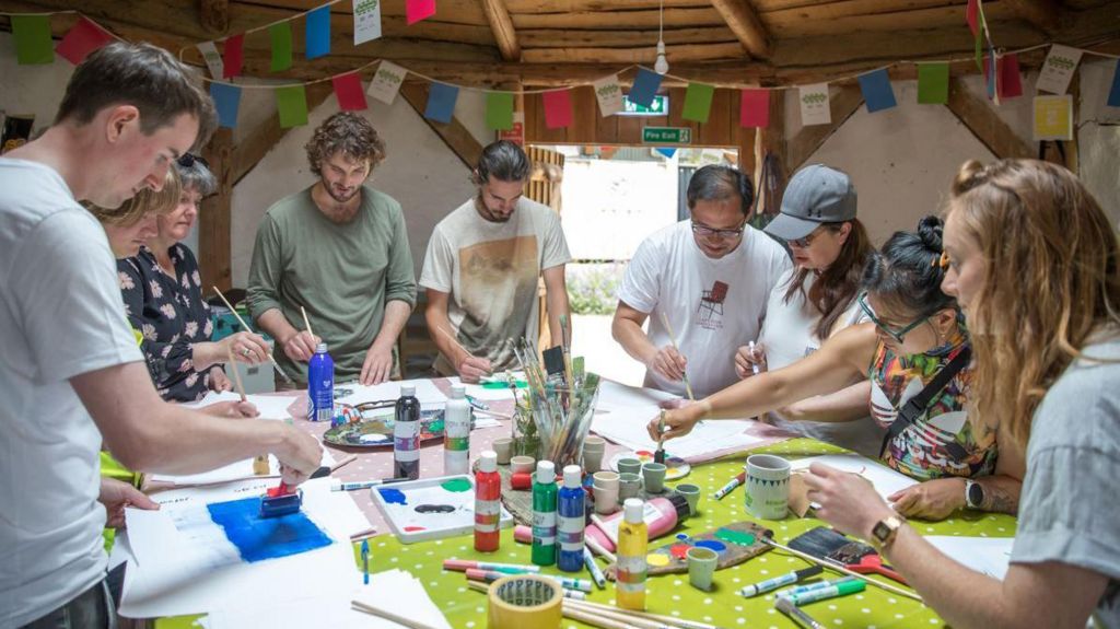 A group of nine people gathered round a table painting. There are art supplies piled in the middle of the table