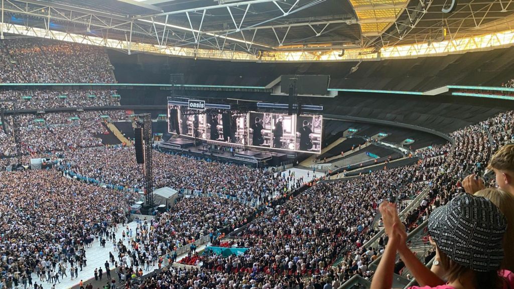 A view from the top tier at Wembley Stadium of the crowd and stage at the concert. The word "Oasis" is printed at the top of the stage.
