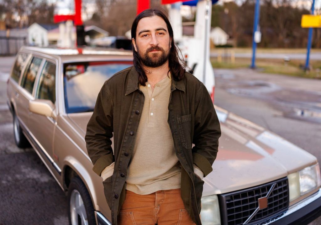 Noah Kahan leans against a vintage station wagon, in a promotional photo for his single The Great Divide.