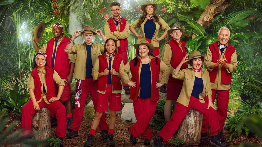 10 people can be seen wearing various outfits of red and khaki green, with some wearing large khaki hats. They are posed in front of a jungle backdrop as part of the announcement for the new cast of I'm a Celebrity...Get Me Out of Here!