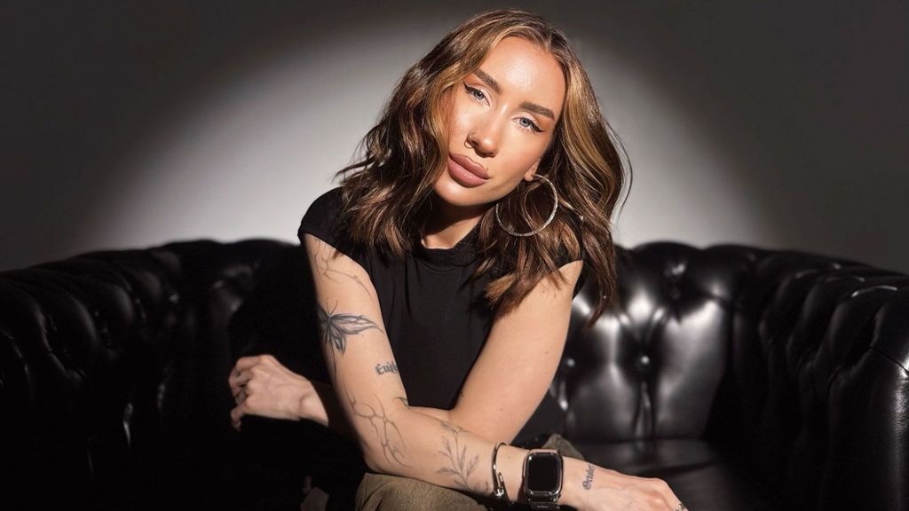 Katya has long mid-brown hair and blue eyes. She is wearing big hoop earrings and has her nose pierced. She has tattooes in the shape of plants and butterflies down one arm.