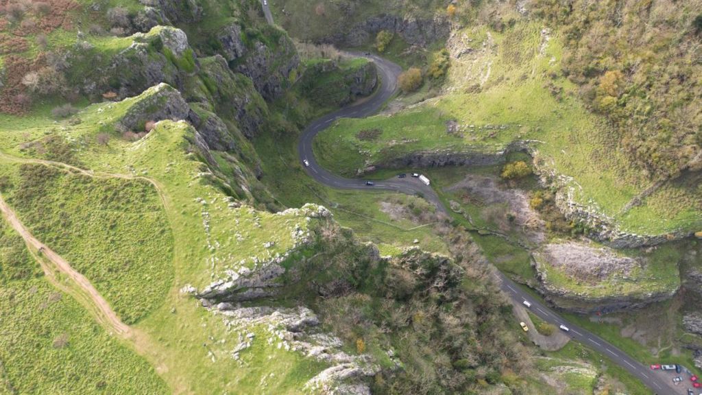 An aerial image of Cheddar Gorge with a long winding road going through it with several cars on it. It is a large rugged landscape with rocks and shrubbery