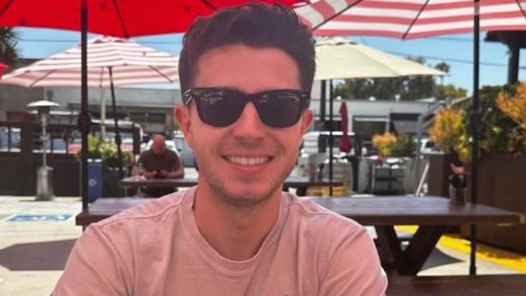 Mason Leposavic smiled while wearing sunglasses, sitting in a spot with picnic tables and umbrellas