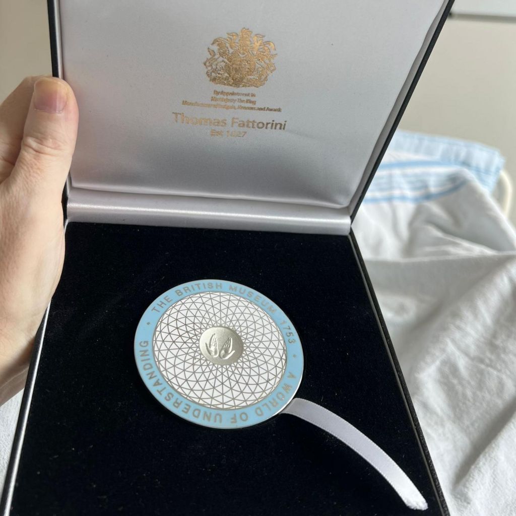 The medal awarded to Dr Ittai Gradel in a presentation box - it is circular, pale blue on the outside with the centre in white and gold, similar in design to the roof of the extension to the British Museum - curved lattice work
