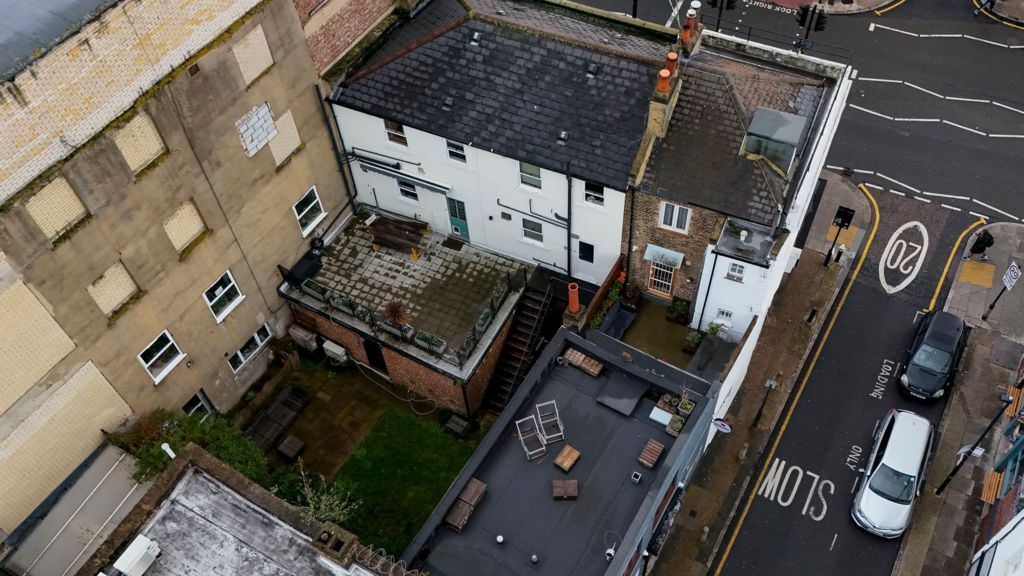 An aerial view of the shared back garden of two neighbouring properties on Bethnal Green Road, east London. It shows a road in front and to the left with cars on it. A roof area with chairs and seating on it, and a small green patch in a garden. Building are around the garden area. 