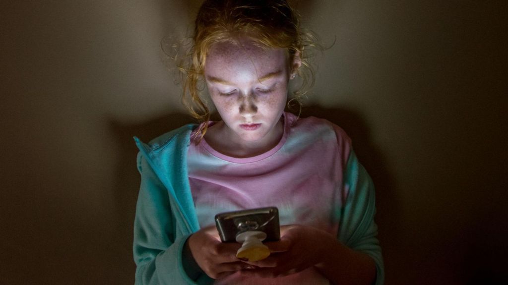 A young long haired redhead girl with face lit up looking into mobile phone screen in a dark room, against a flat wall, with sad facial expression