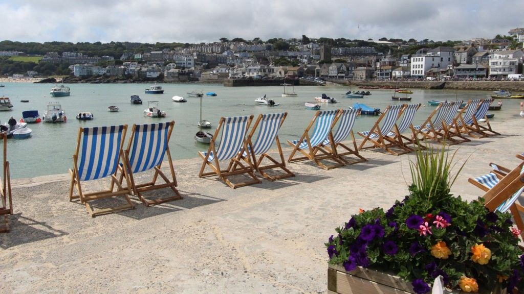 A row of empty blue and white deck chairs look out over a harbour full of boats with houses on the other side