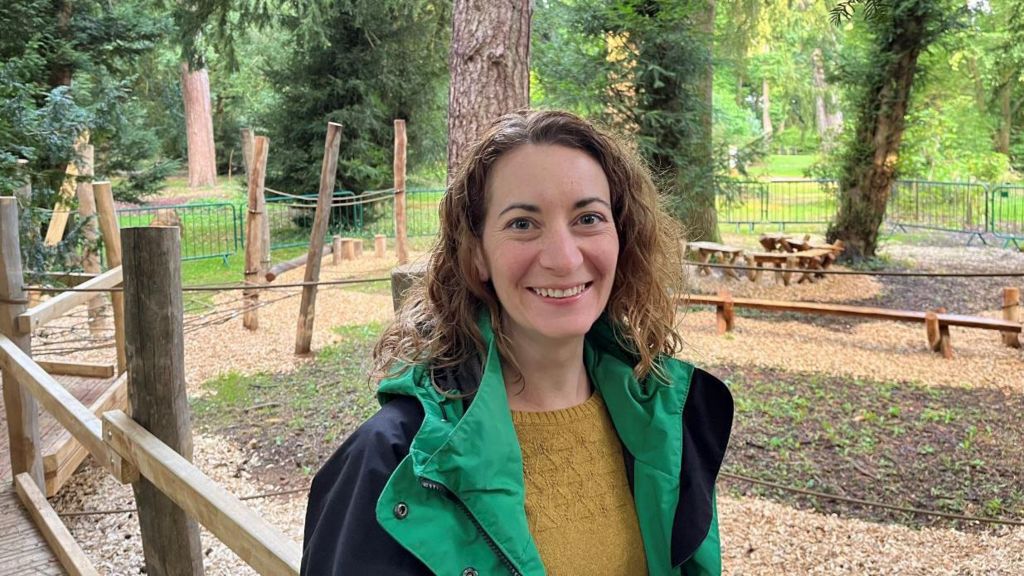 Sophie Nash from Forestry England wearing a green and black raincoat and yellow jumper. She has shoulder length wavy hair and is smiling at the camera. In the background there are trees, bark chipping paths and wooden climbing frames 