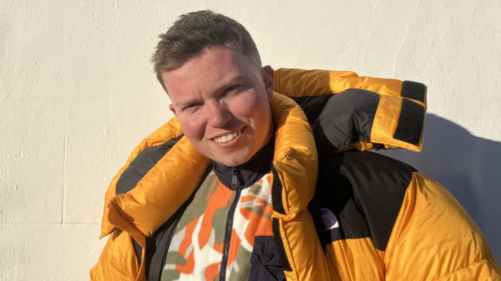A man wearing a bright yellow puffer coat smiles at the camera. He has short brown hair and is wearing a patterned fleece under his coat. He is standing in front of a cream-coloured wall.