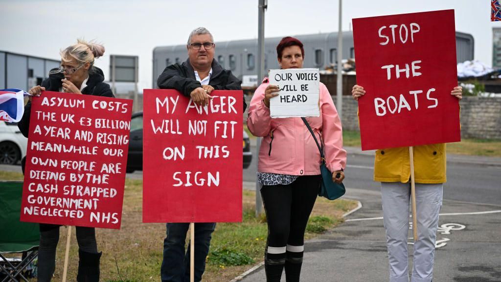 Four people holding red signs. One reads: "Asylum costs the UK £3 billion a year and rising meanwhile our own people are dying by the cash strapped government neglected NHS." Others read: "My anger will not fit on this sign", "our voices will be heard", and "stop the boats".