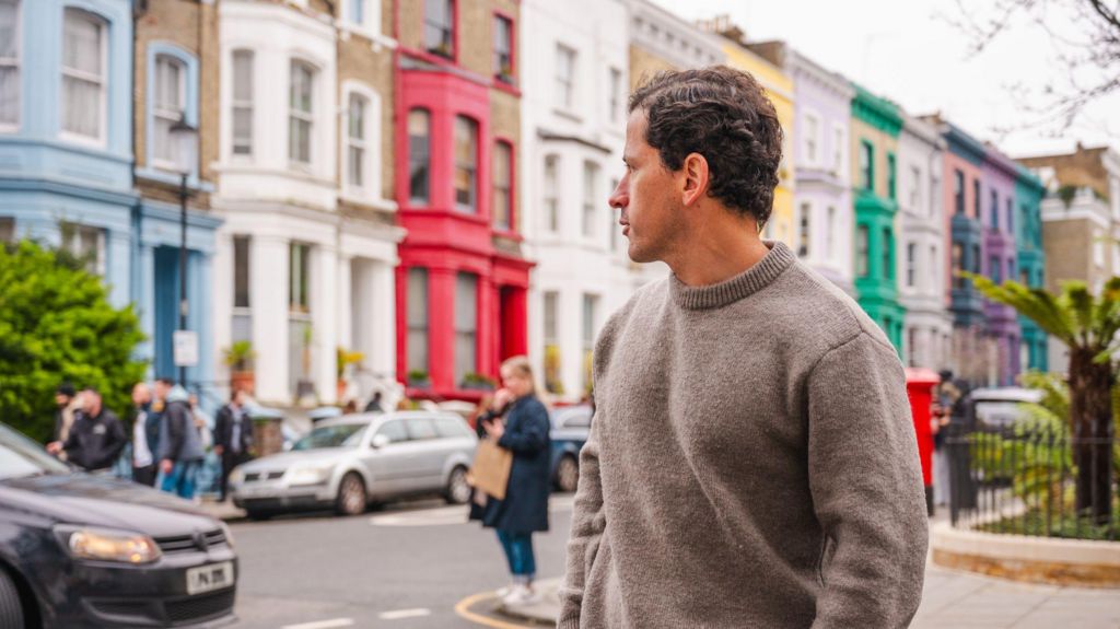 Young man in a jumper looks back over his shoulder at a row of brightly painted terraced Notting Hill townhouses.