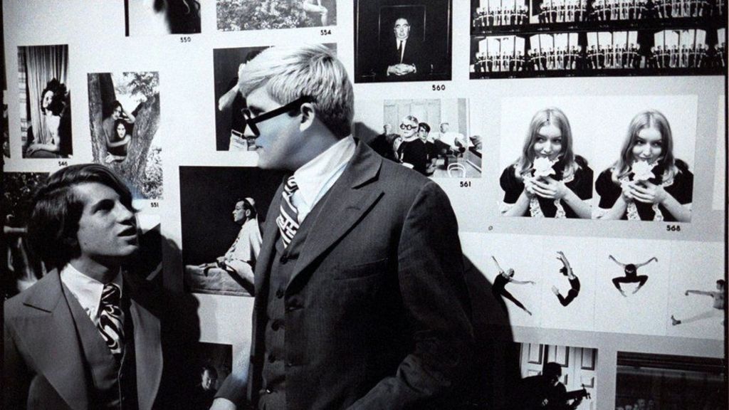 David Hockney and Peter Schlesinger. This is a black and white photo of Hockney and his former boyfriend. They are in a gallery surrounded by black and white photos. They are captured while having a conversation.