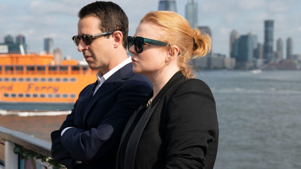 Jeremy Strong and Sarah Snook from Succession stand by the water in sunglasses and suits, with New York City in the background