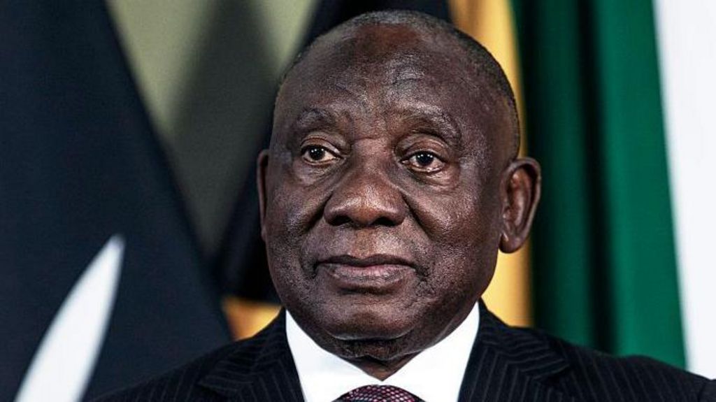 A head-and-shoulders image of Cyril Ramaphosa. He is wearing a dark suit and the white collar of his shirt can just be seen. Some flags are out of focus in the background.