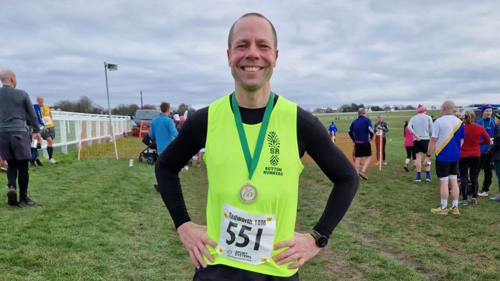 Wood wearing a yellow running vest, with a running track and other runners in the background, resting his hands on his hips, smiling into the camera. He is wearing a medal on a green strap around his neck.
