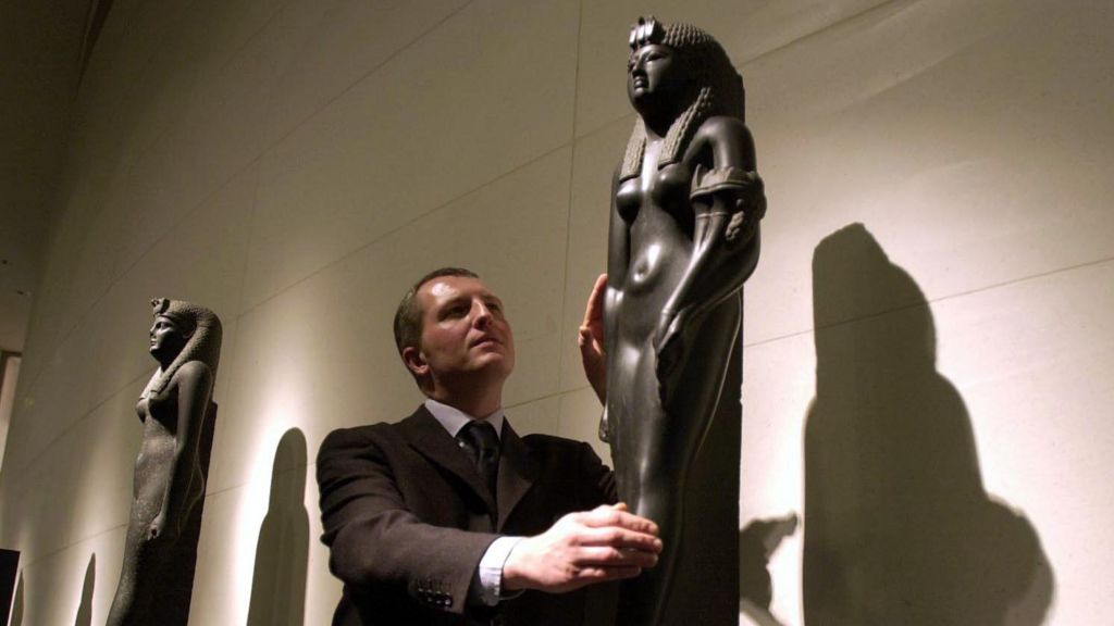 Dr Peter Higgs in brown suit in centre of image with a black basalt statue of Cleopatra VII,  at the British Museum in 2001
Another statue of Cleopatra behind him.