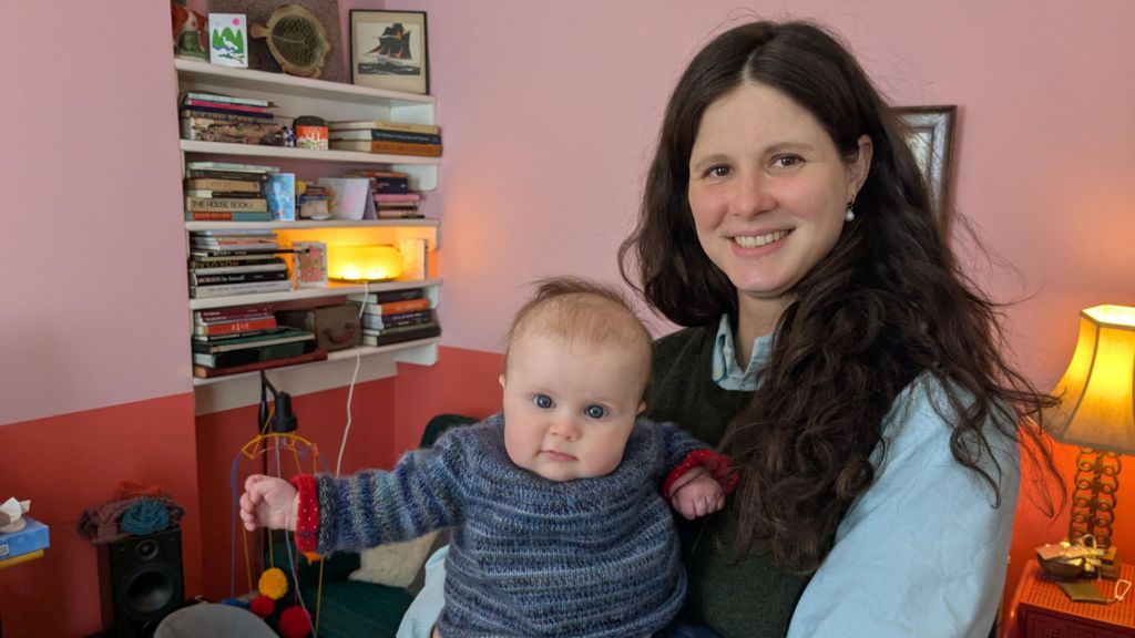 Georgie Scott, a woman with long brown hair in a blue shirt and jumper waistcoat holds a baby in a living room. The baby smiles into the camera, and is wearing a knitted blue jumper. 