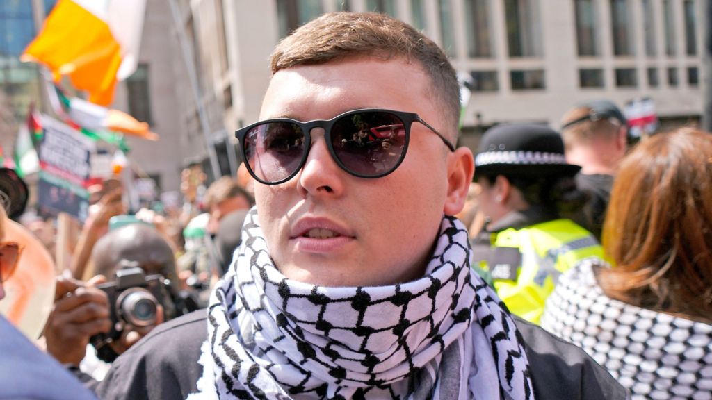 An up close image of Liam Óg Ó hAnnaidh, who is wearing a scarf and sunglasses. There is a crowd of people behind him.