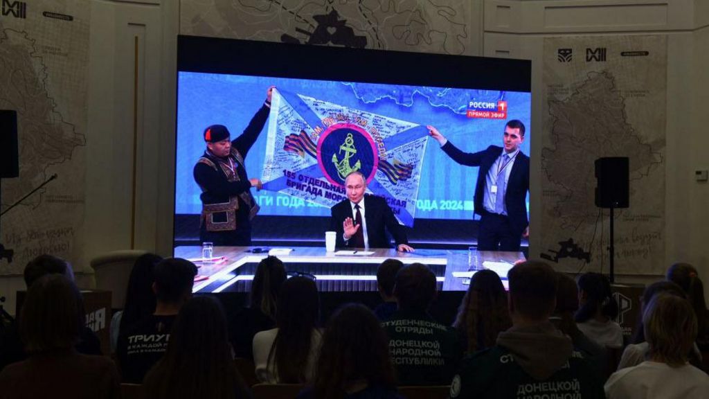 Putin on a TV screen, behind him staff members hold-up a flag he said was given to him by Russian marines fighting in Kursk