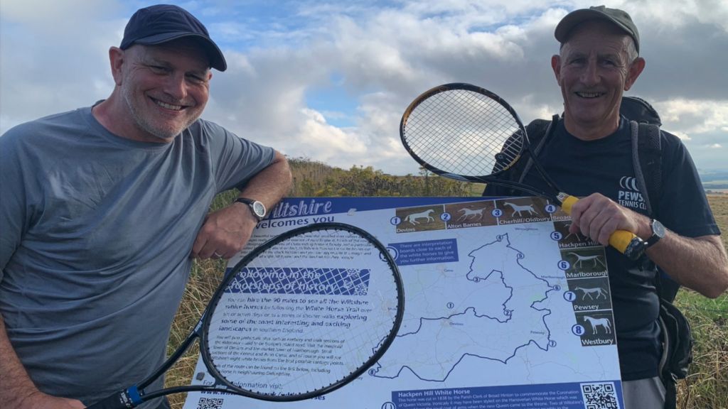 Two men smiling at the camera and holding tennis rackets, while posing on an information board about the walk