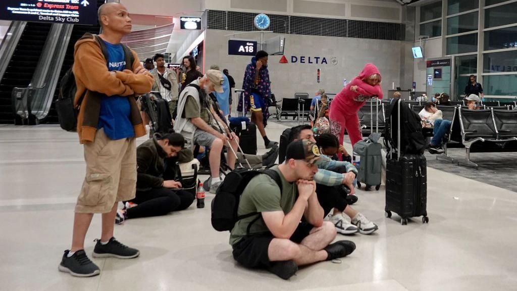 A group of people sit on the floor in an airport while waiting for a flight. They all look bored and exhausted.