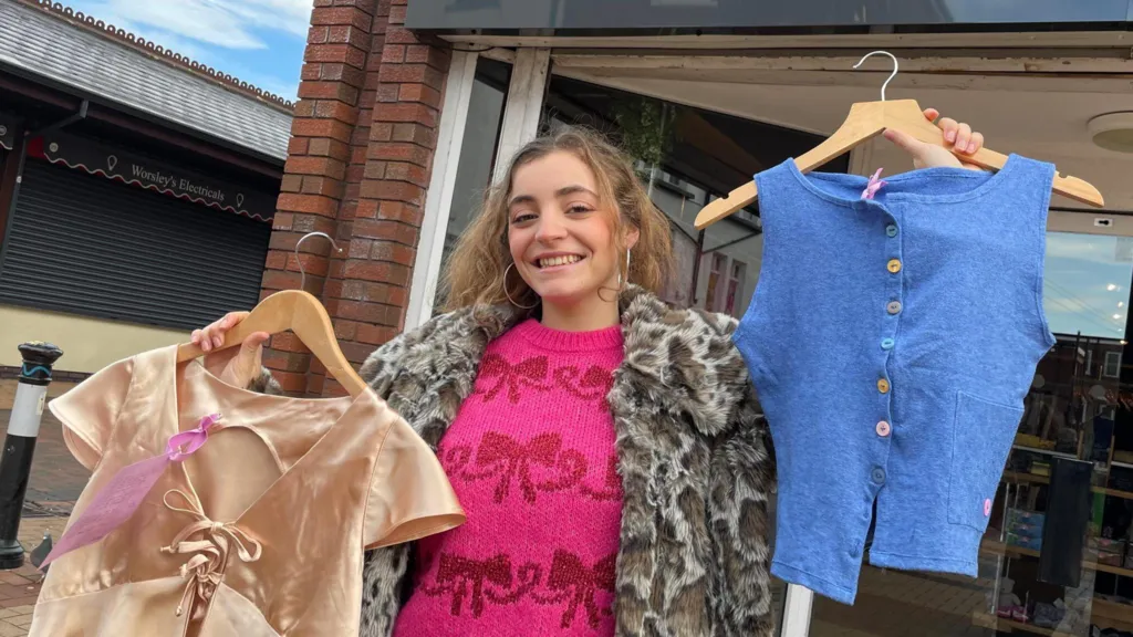 Fashion designer Victoria Ford with long brown wavy hair wearing large silver hooped earrings and a faux fur leopard skin coat and hot pink knitted jumper with dark red bows on it holds two of her designs up outside a charity shop. In her right hand (pictured left) is a gold ladies top on a coat hanger and in her left hand she holds up a blue sleeveless top on a coat hanger. She is smiling. 