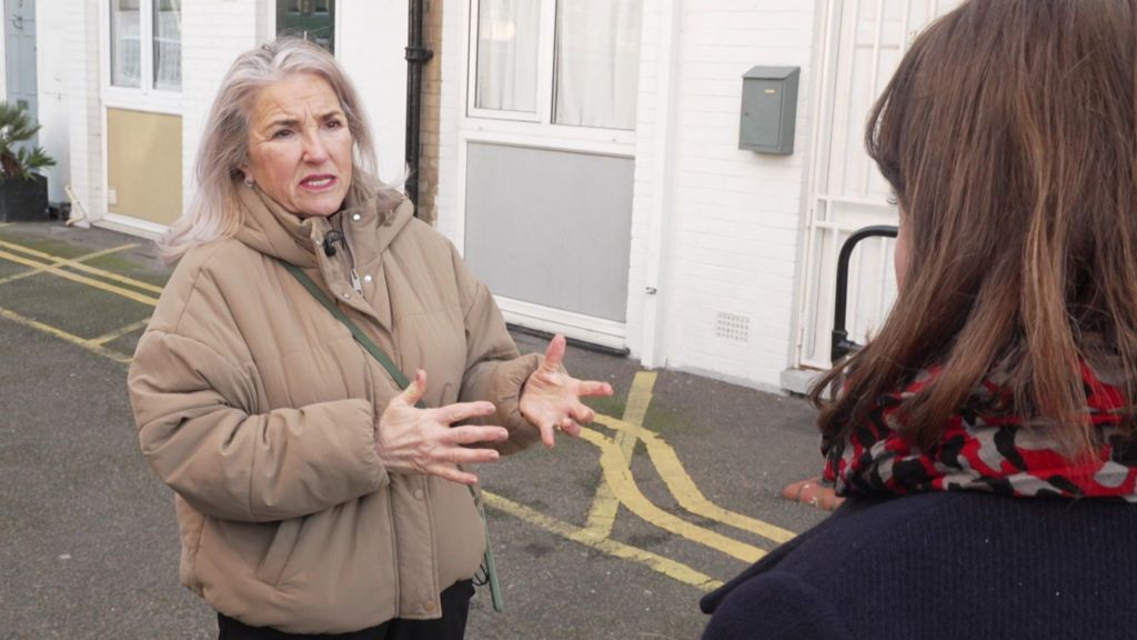An older lady with long grey hair talks to another woman outside some flats in east London