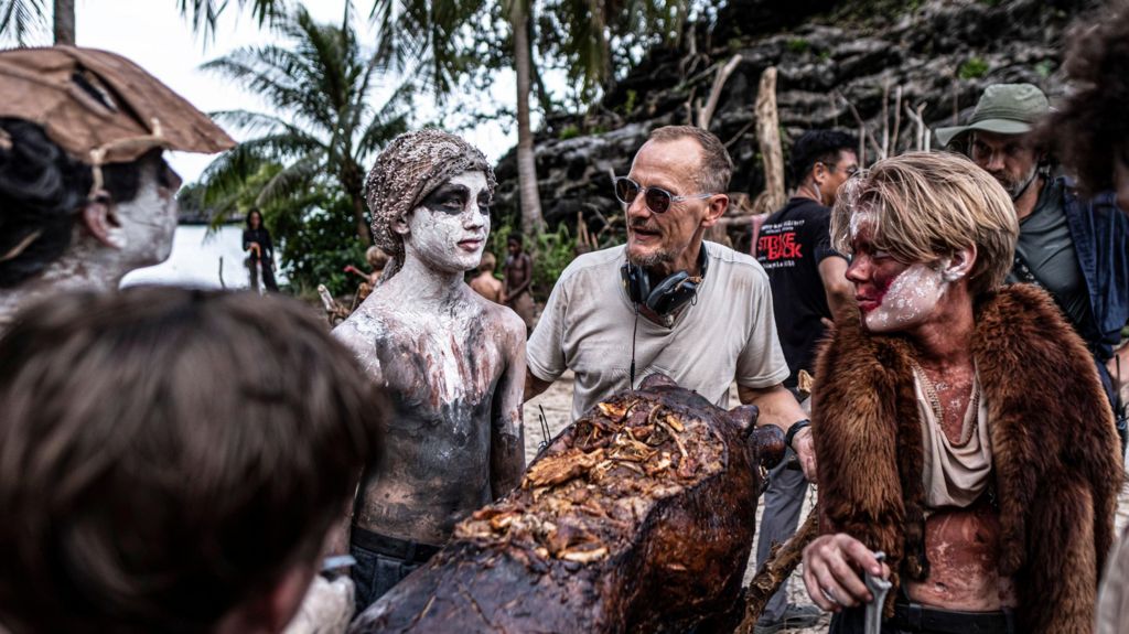 Marc Munden, wearing sunglasses with headphones around his neck, speaks to two of the actors, who are in costume.