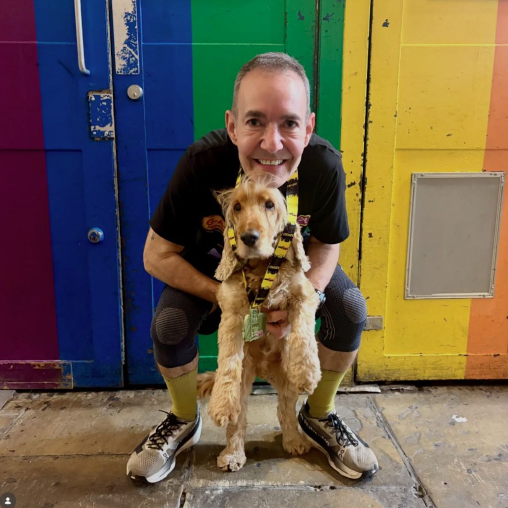 A man dressed in running gear and a big smile crouches with his dog against a rainbow wall.