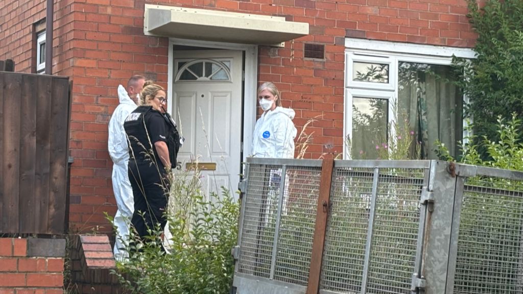 A police officer and two other people in white suits, one in a mask, at the open door of a house